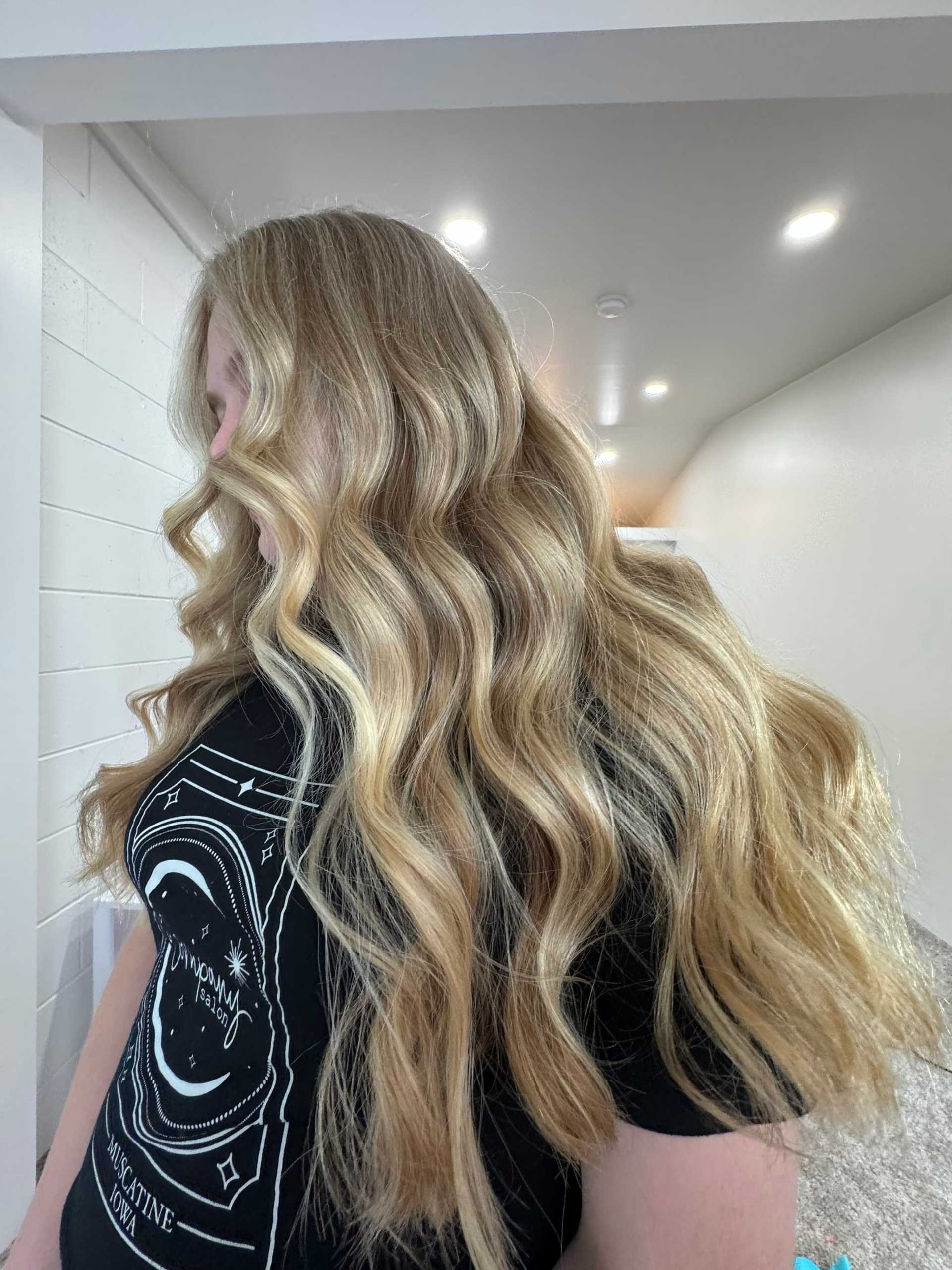 Person with long, wavy blonde hair standing in a well-lit hallway, wearing a black graphic T-shirt.